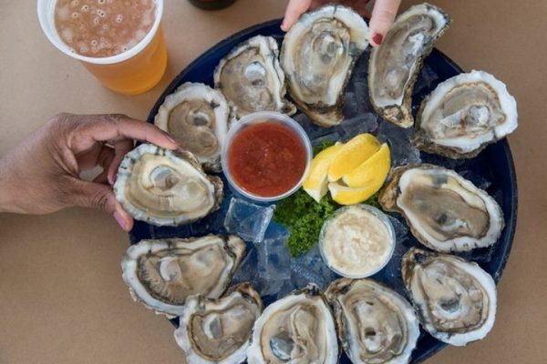 close up of oysters on platter with hands holding two of them and beers